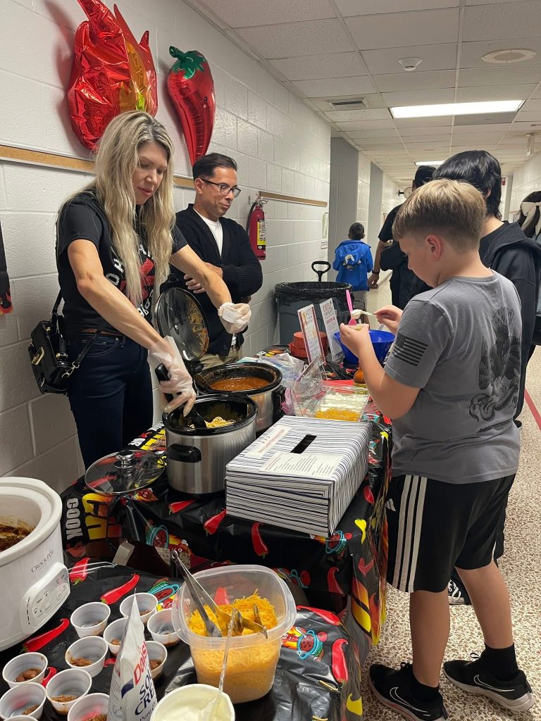 Adult volunteers serving chili to a child at a kindergarten chili cook-off.
