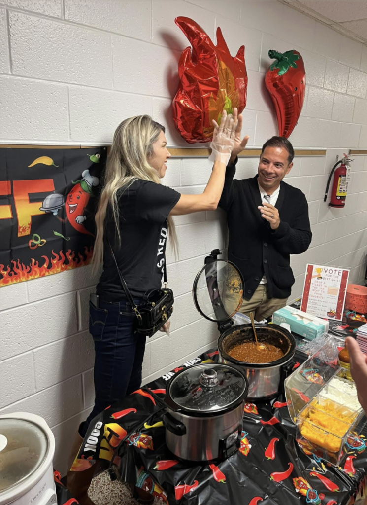 Adult volunteers high-fiving while serving chili at a kindergarten chili cook-off table.