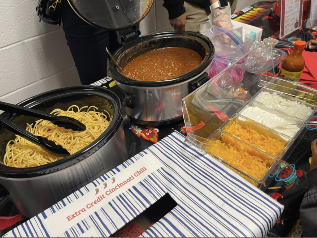 Chili cook-off serving table with slow cookers, spaghetti, shredded cheese, sour cream, and toppings.